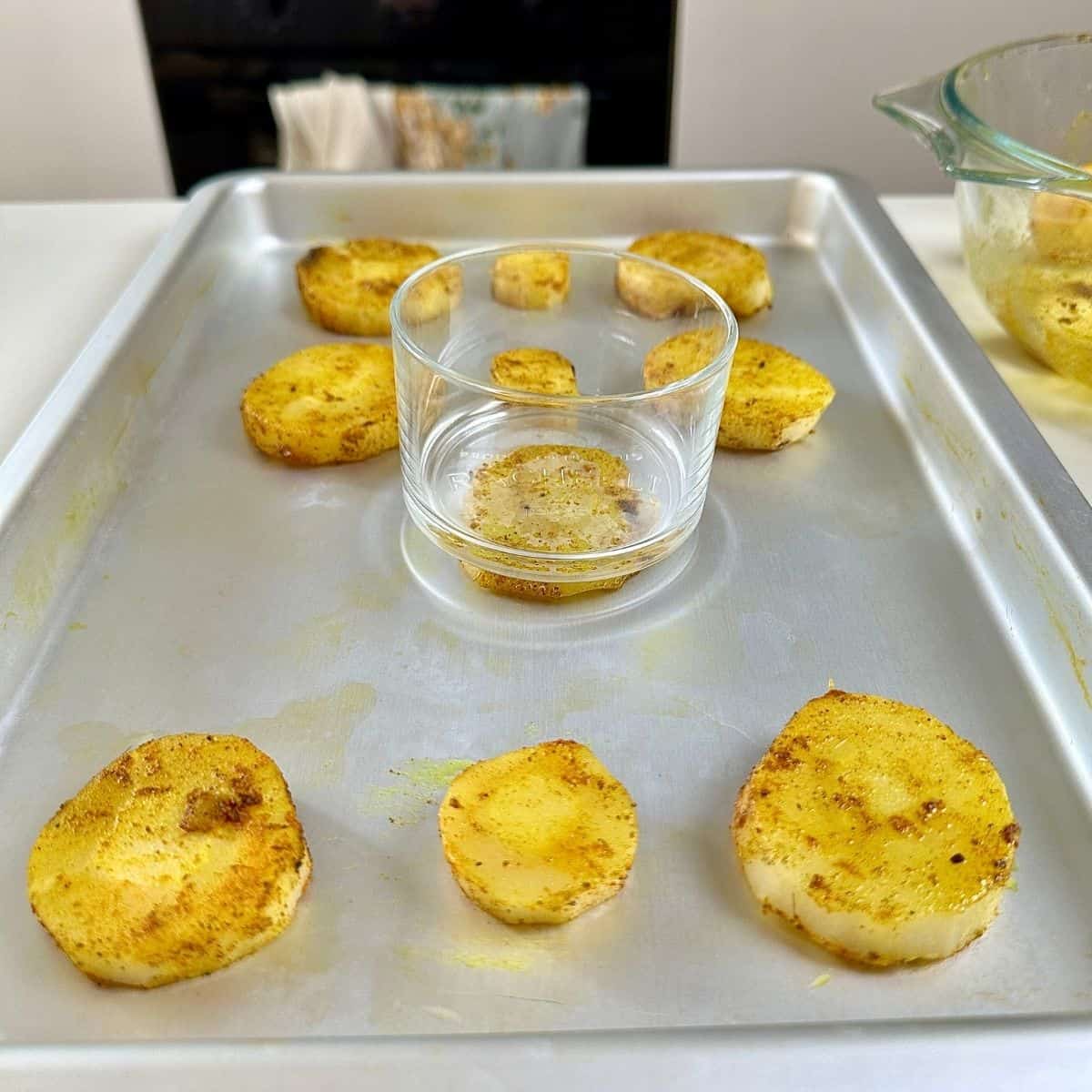 Parsnip slices coated with olive oil, cumin powder and turmeric powder on a baking tray. A flat-bottomed glass sits on one of the slices that phas been pressed down by the glass, creating a "smashed" parsnip slice.