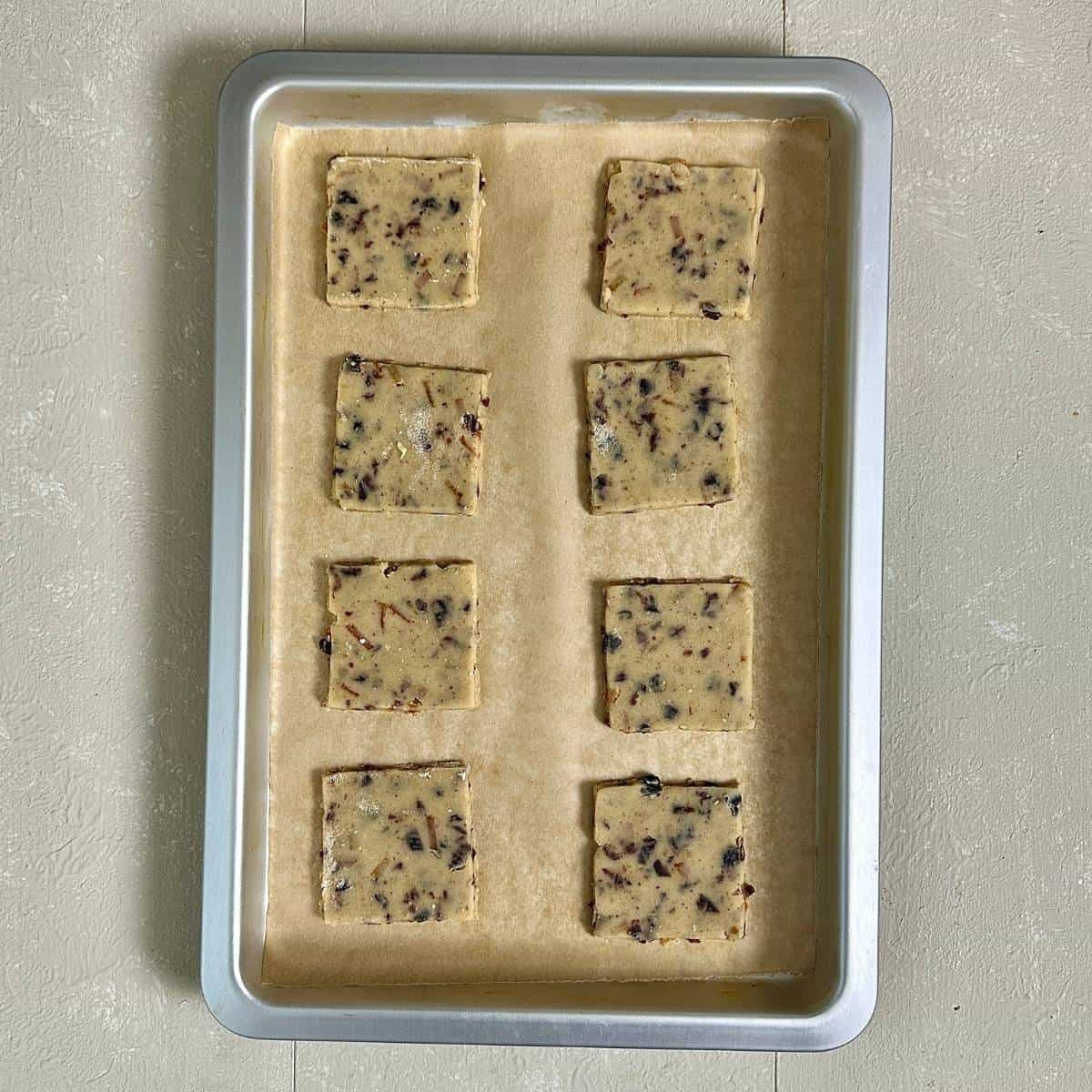 A silver baking tray lined with parchment paper containing eight square vegan Christmas biscuits, ready for baking.