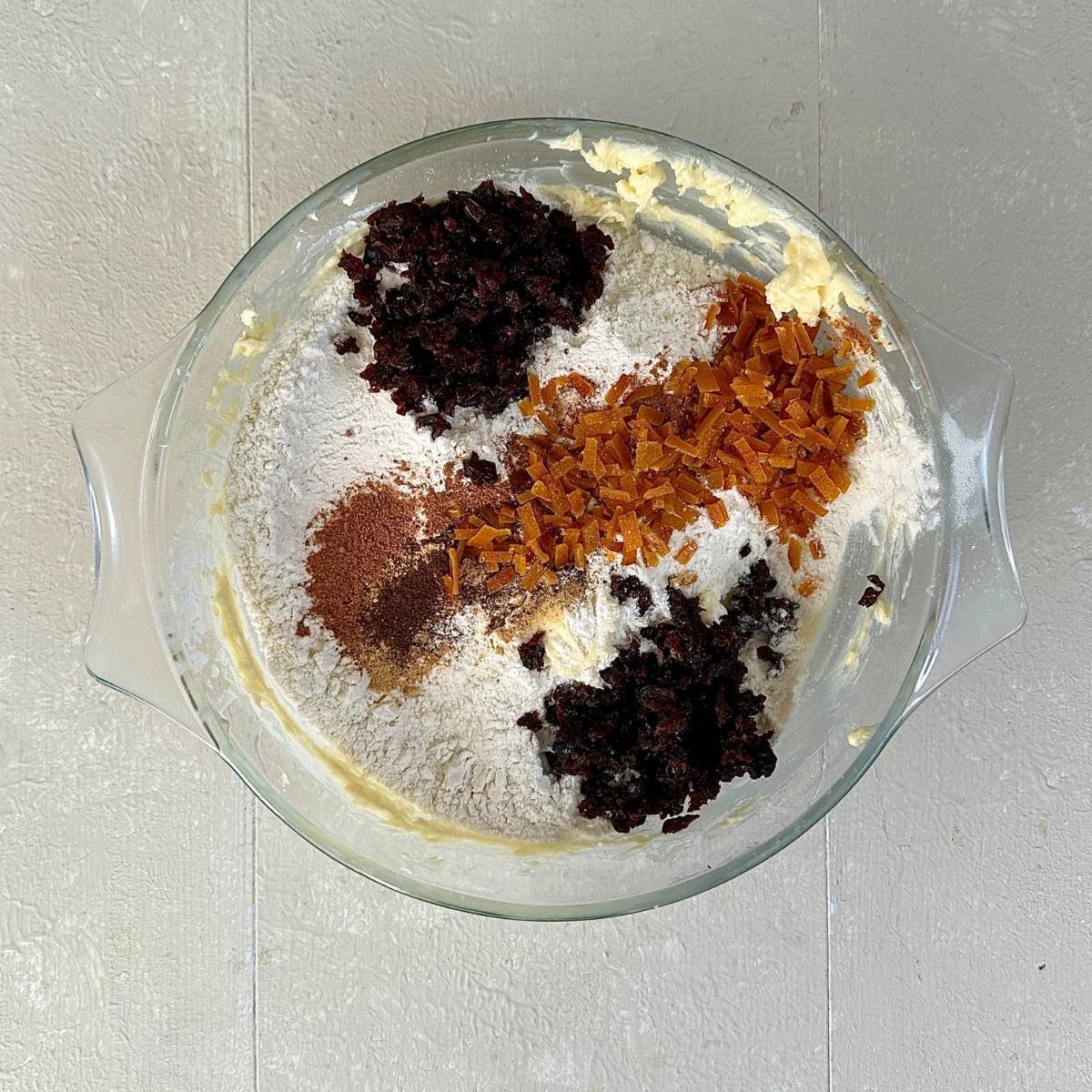 A glass bowl containing spices, chopped dried fruit and flour ready to combine into creamed icing sugar and butter, to make a dough for vegan Christmas biscuits.