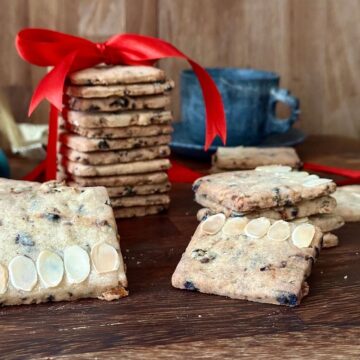 Square-shaped vegan Christmas biscuits. A tower of biscuits wrapped in a red ribbon sits in the background.