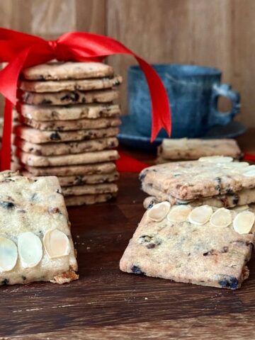 Square-shaped vegan Christmas biscuits. A tower of biscuits wrapped in a red ribbon sits in the background.