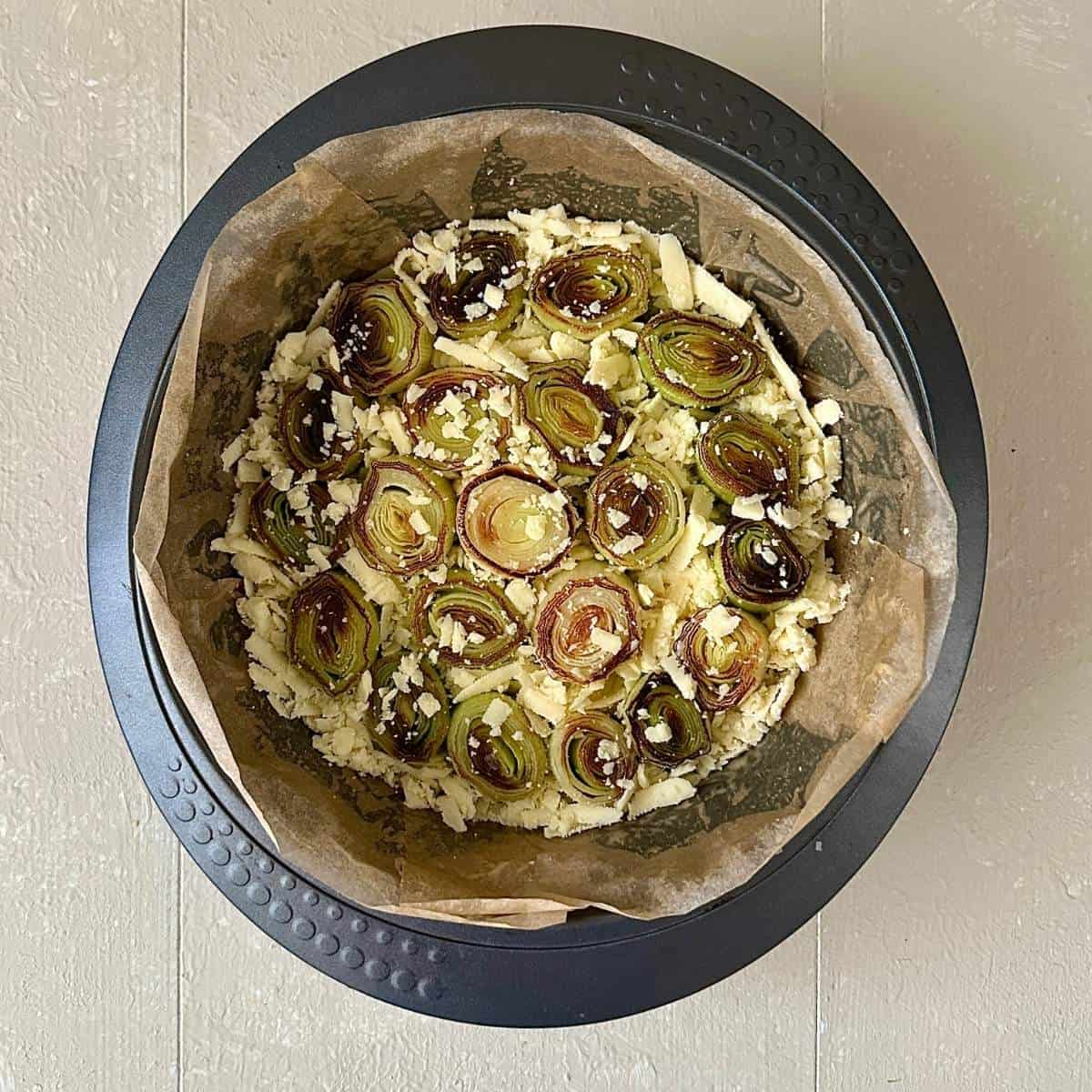 A round cake tin lined with parchment paper containing ยพ inch thick leek lengths arranged in the bottom of the tin with grated vegan cheddar filling the gaps. Part of a vegan christmas bake.