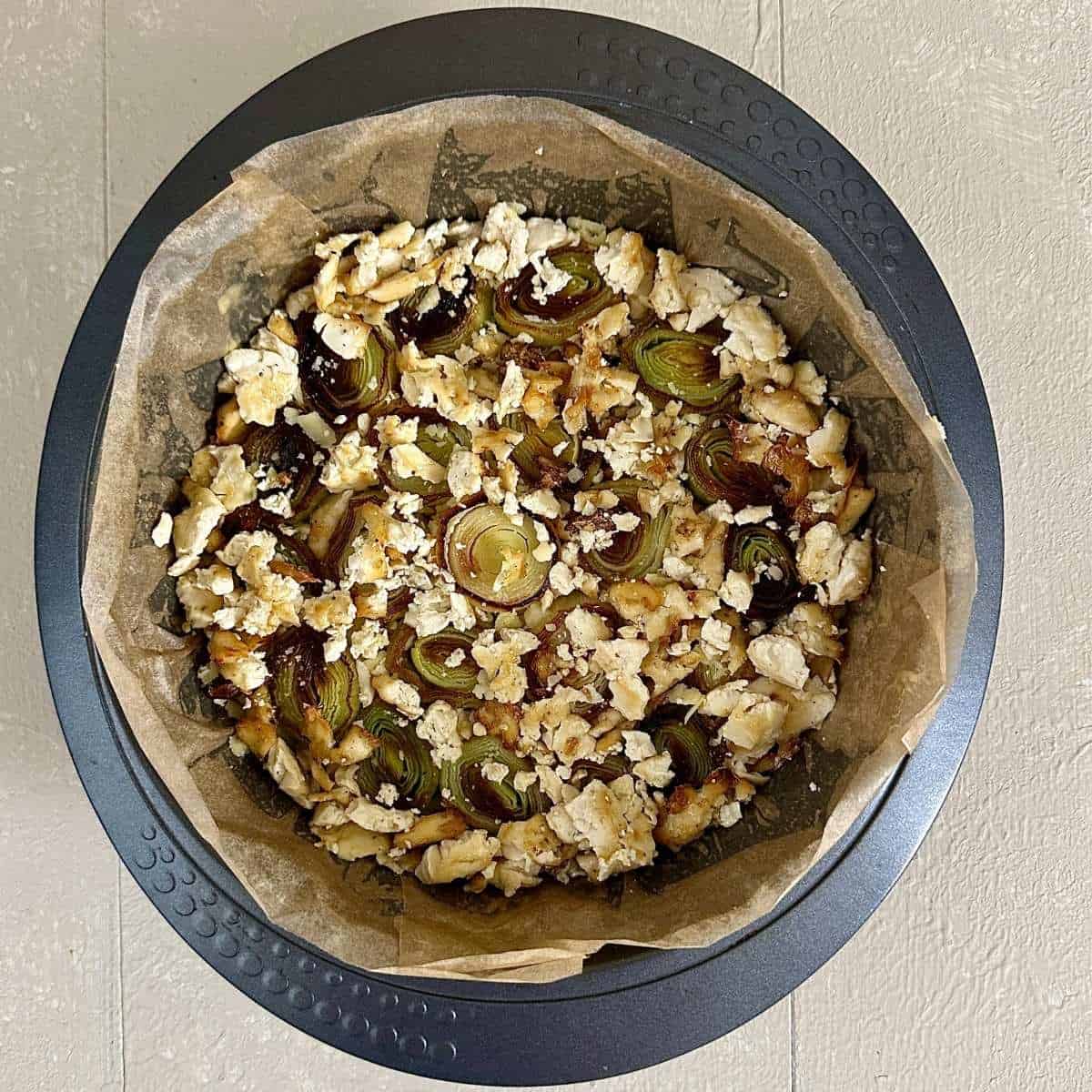 A large cake tin lined with parchment paper containing layered ingredients for a vegan Christmas bake. A layer of ยพ inch leek lengths with vegan cheddar filling the gaps between the leek chunks and a layer of cooked firm tofu chunks on top.
