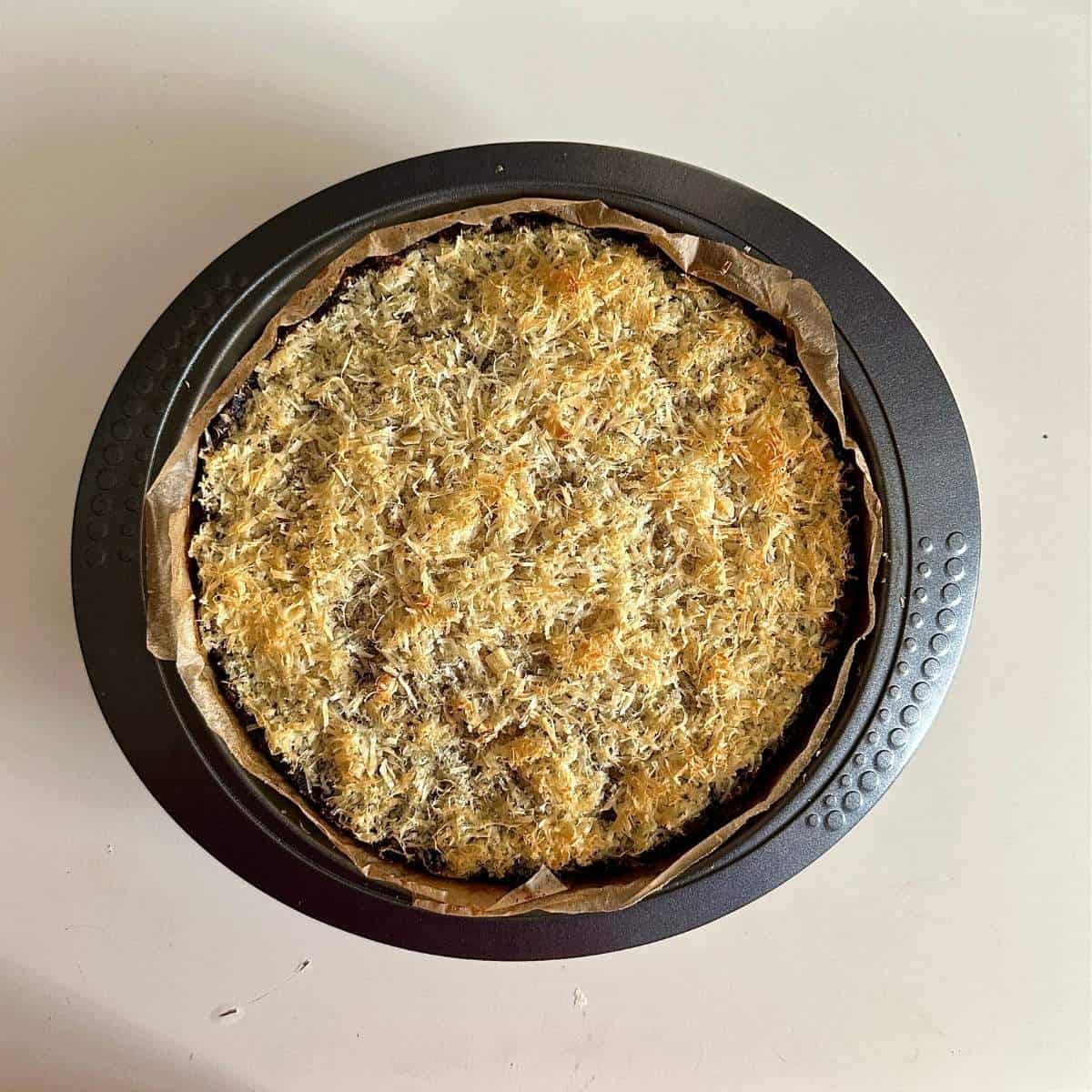 A large round cake tin containing a vegan Christmas bake after cooking. A layer of grated vegan parmesan cheese browned under a grill lays level with the top of the tin.
