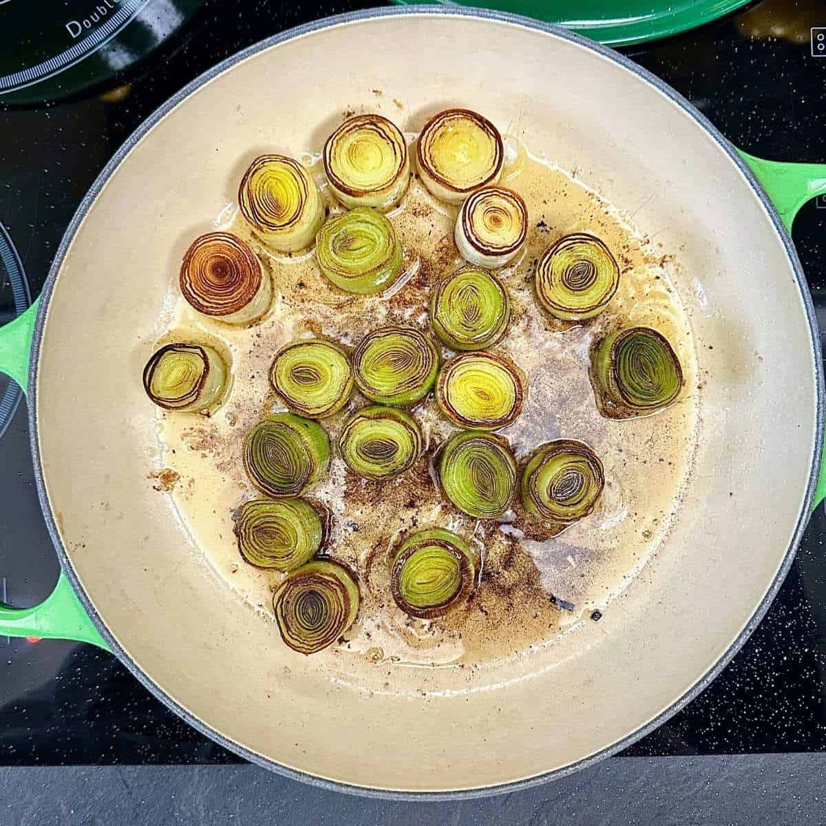 A large green saucepan containing thickly cut lengths of leek with caramelised ends, sitting in olive oil and melted butter.