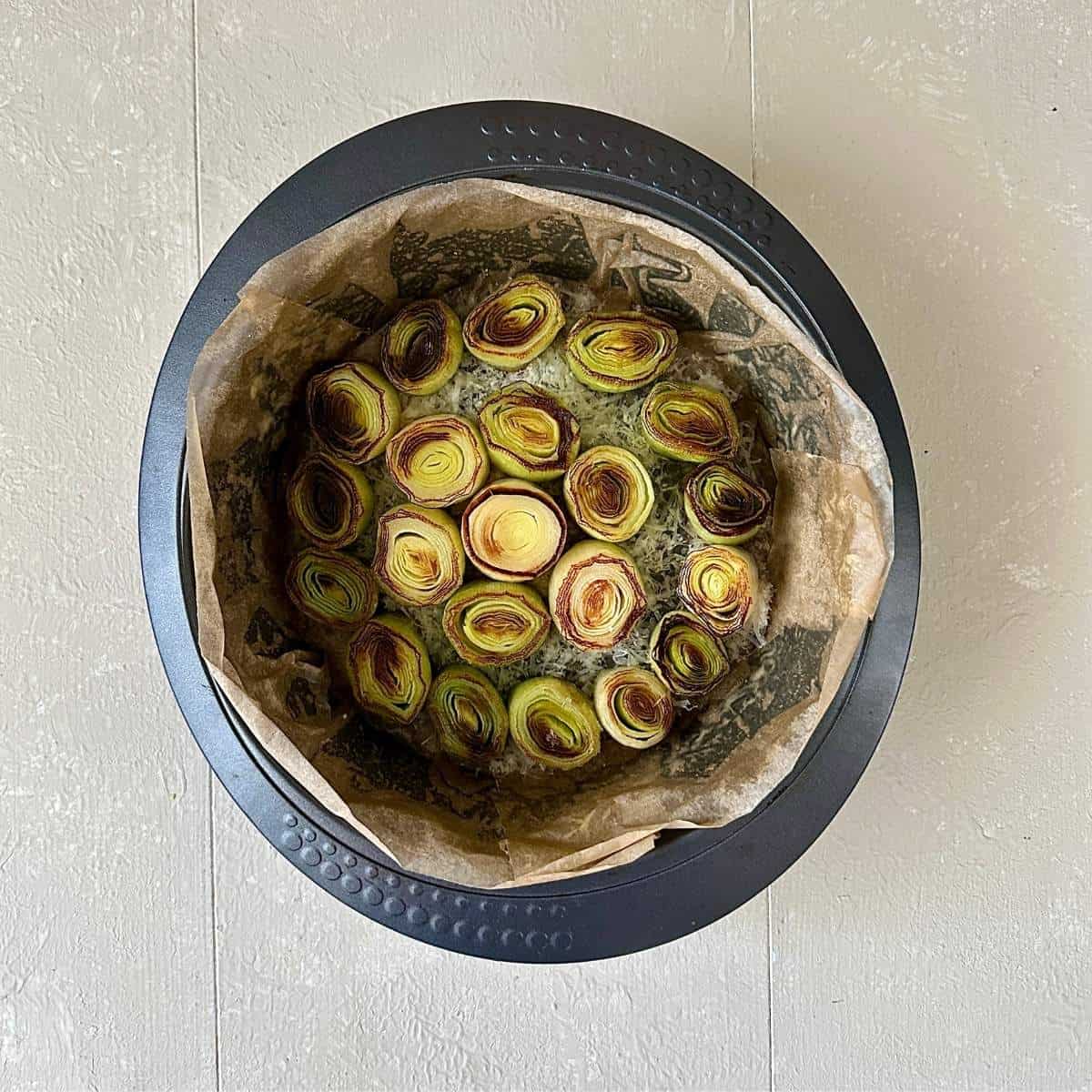 A large round cake tin lined with parchment paper containing a layer of grated parmesan cheese with thick slices of caramelised leek sitting on top.