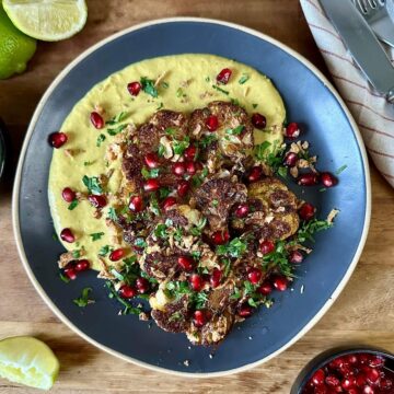 A blue ceramic plate with roasted and caramelised cauliflower steaks and a smooth butterbean korma-style sauce. Garnished with chopped coriander and pomegranate seeds.