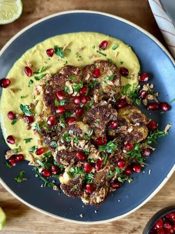 A blue ceramic plate with roasted and caramelised cauliflower steaks and a smooth butterbean korma-style sauce. Garnished with chopped coriander and pomegranate seeds.