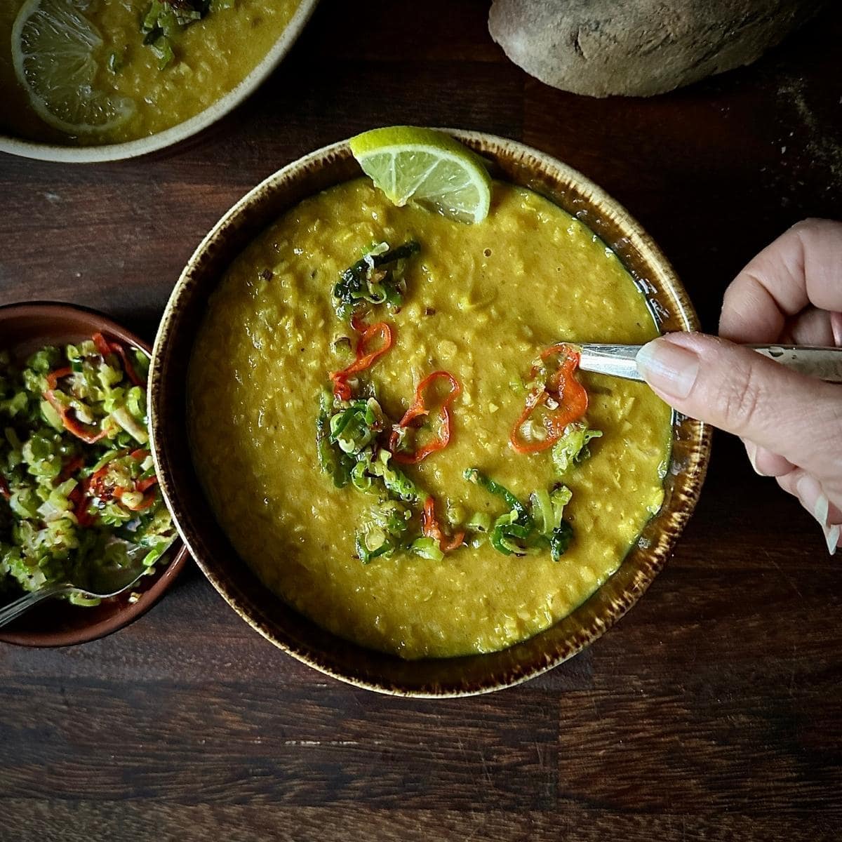 A bowl of leek and lentil soup topped with chilli and leeks, with a lime wedge on the rim and a hand reaching in with a spoon - cosy comforting cold weather soup.