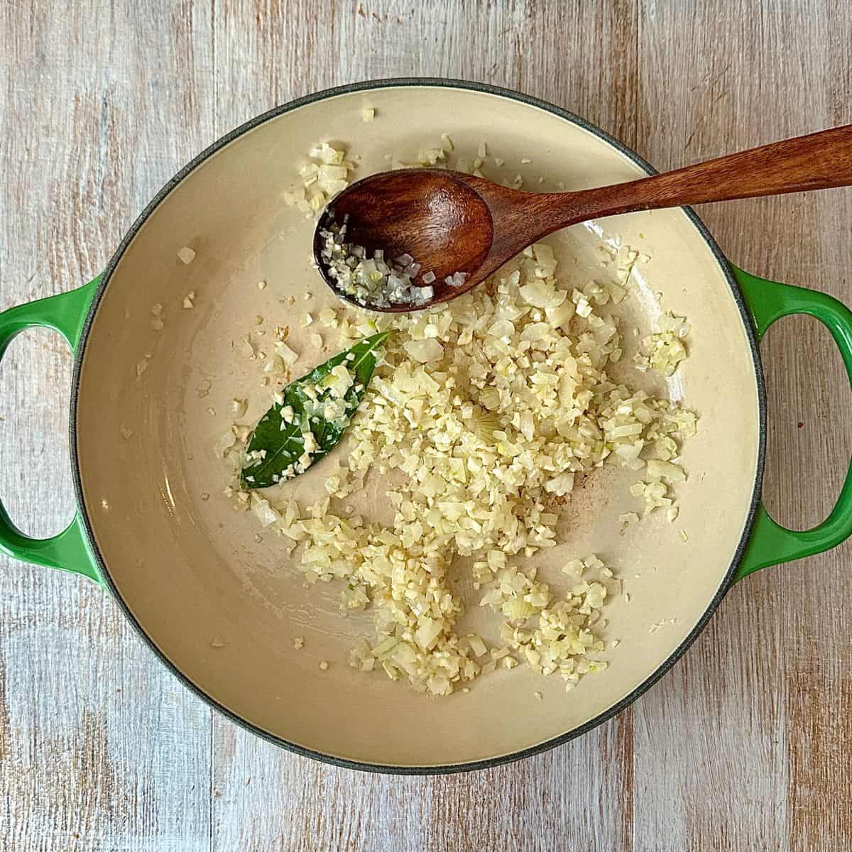 A large green pot containing finely chopped white onion and garlic and a bay leaf for pumpkin casserole recipe. A wooden spoon sits in the pot.