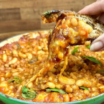 Close up of a piece of toast covered in cheesy pizza beans being held over a large pan containing cheesy pizza beans.