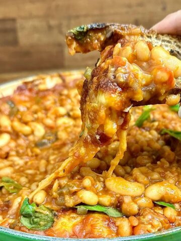 Close up of a piece of toast covered in cheesy pizza beans being held over a large pan containing cheesy pizza beans.