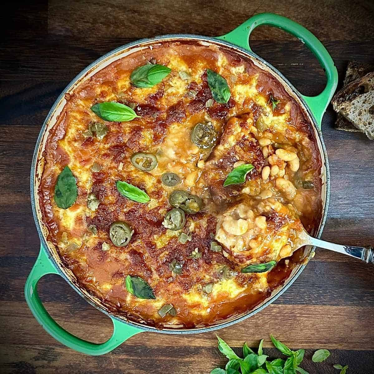 A large green pan containing pizza beans cooked and ready to serve. A spoon with a scoop of pizza beans in help above the right hand side of the pan.