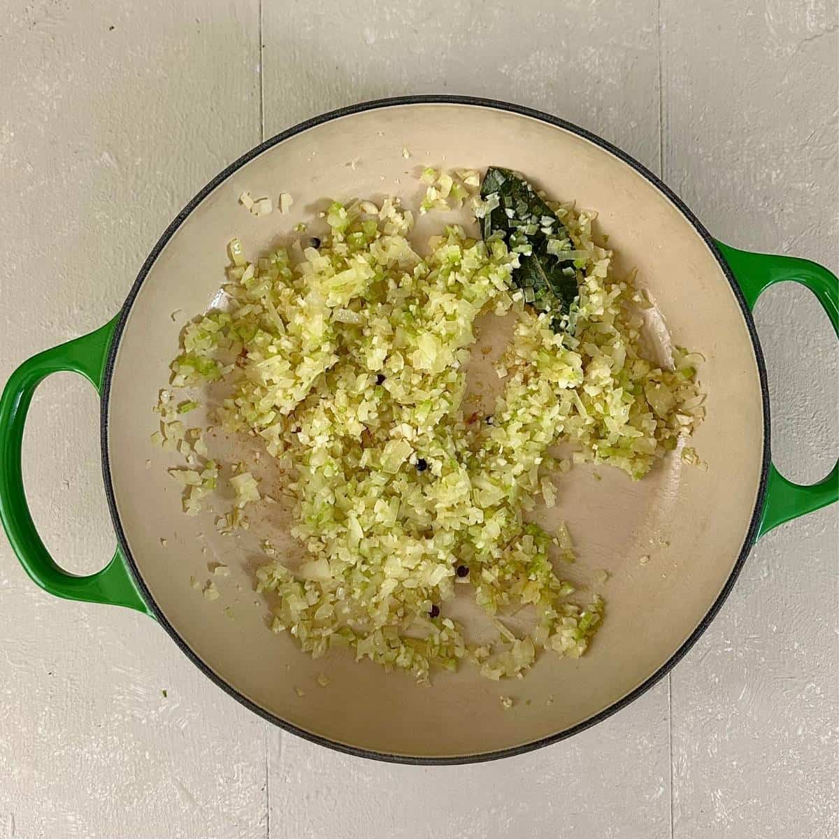 A large green pot containing finely chopped onion, garlic, celery, peppercorns and a bayleaf, for cheesy pizza beans recipe.