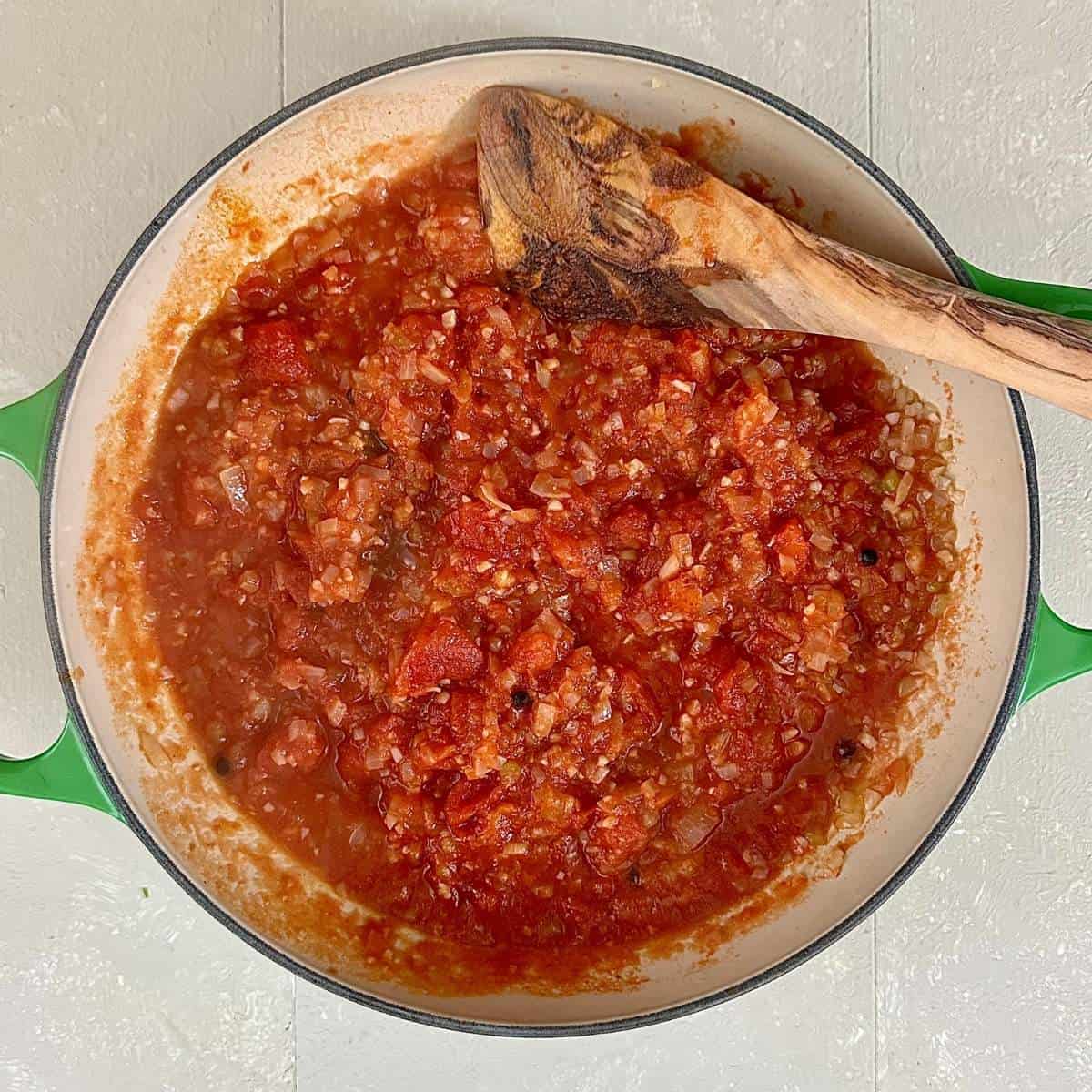 A large green shallow pot containing chopped tinned tomatoes will finely chopped onion, garlic and celery, peppercorns and bay leaf for cheesy pizza beans recipe.