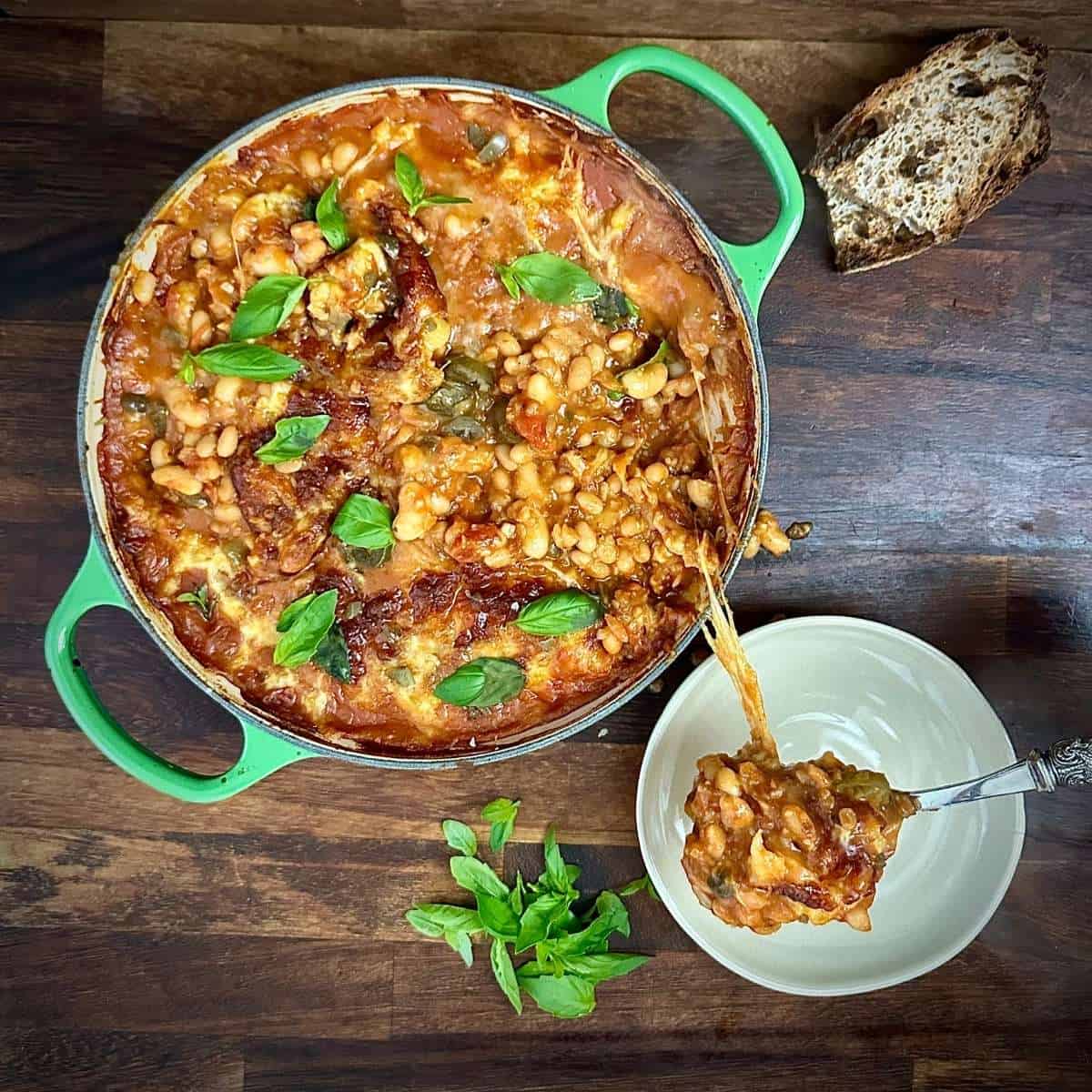 A large green pan containing pizza beans with a smaller dish next to it containing a spoon with pizza beans on it, with cheese pull from the main pan.