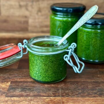 A small glass jar containing a vivid green herb sauce. A spoon sits in the jar. Two more jars of herb sauce sit in the background.