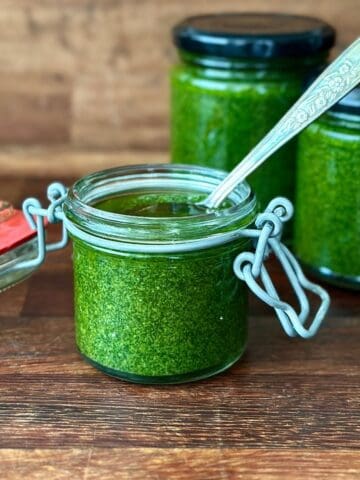A small glass jar containing a vivid green herb sauce. A spoon sits in the jar. Two more jars of herb sauce sit in the background.