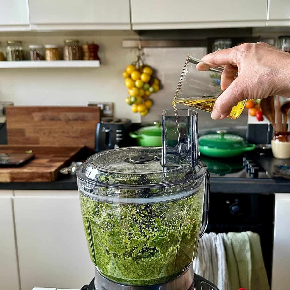 An electric blender containing herb sauce ingredients. The machine is blending and olive oil is being poured into the blender jug.