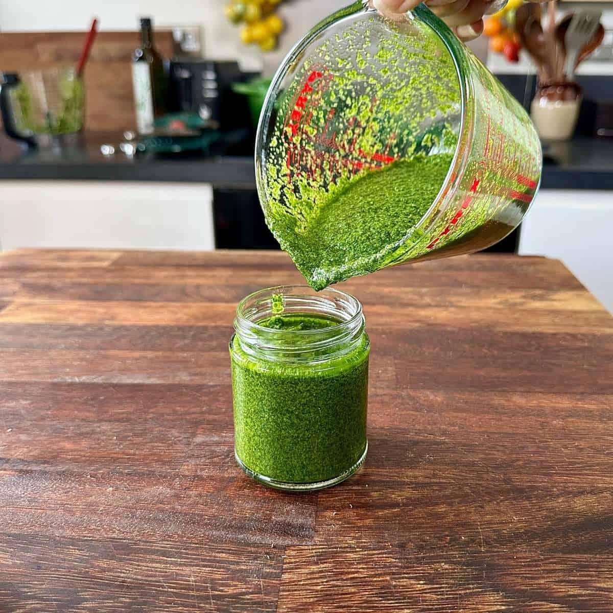 A small jar containing herb sauce. The Sauce is being poured into the jar from a glass measuring jug.