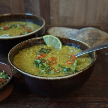 A rustic bowl of leek and lentil soup seen from the side, topped with jalapeรฑo peppers and spring onion, with bread and another bowl in the background.
