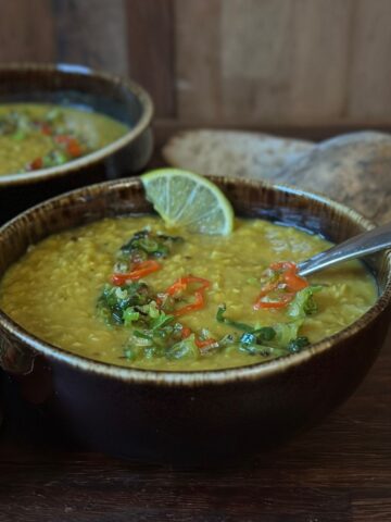A rustic bowl of leek and lentil soup seen from the side, topped with jalapeño peppers and spring onion, with bread and another bowl in the background.