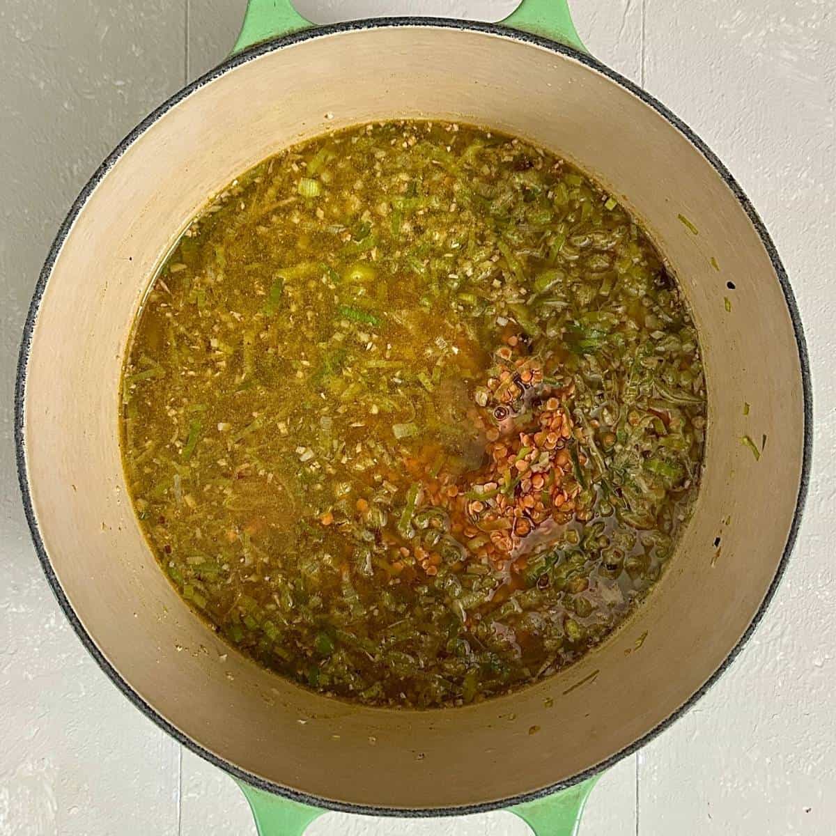 A large green pot containing ingredients for leek and lentil soup: sliced leek, red lentils, chopped ginger and garlic, and vegetable stock. Ready to simmer.