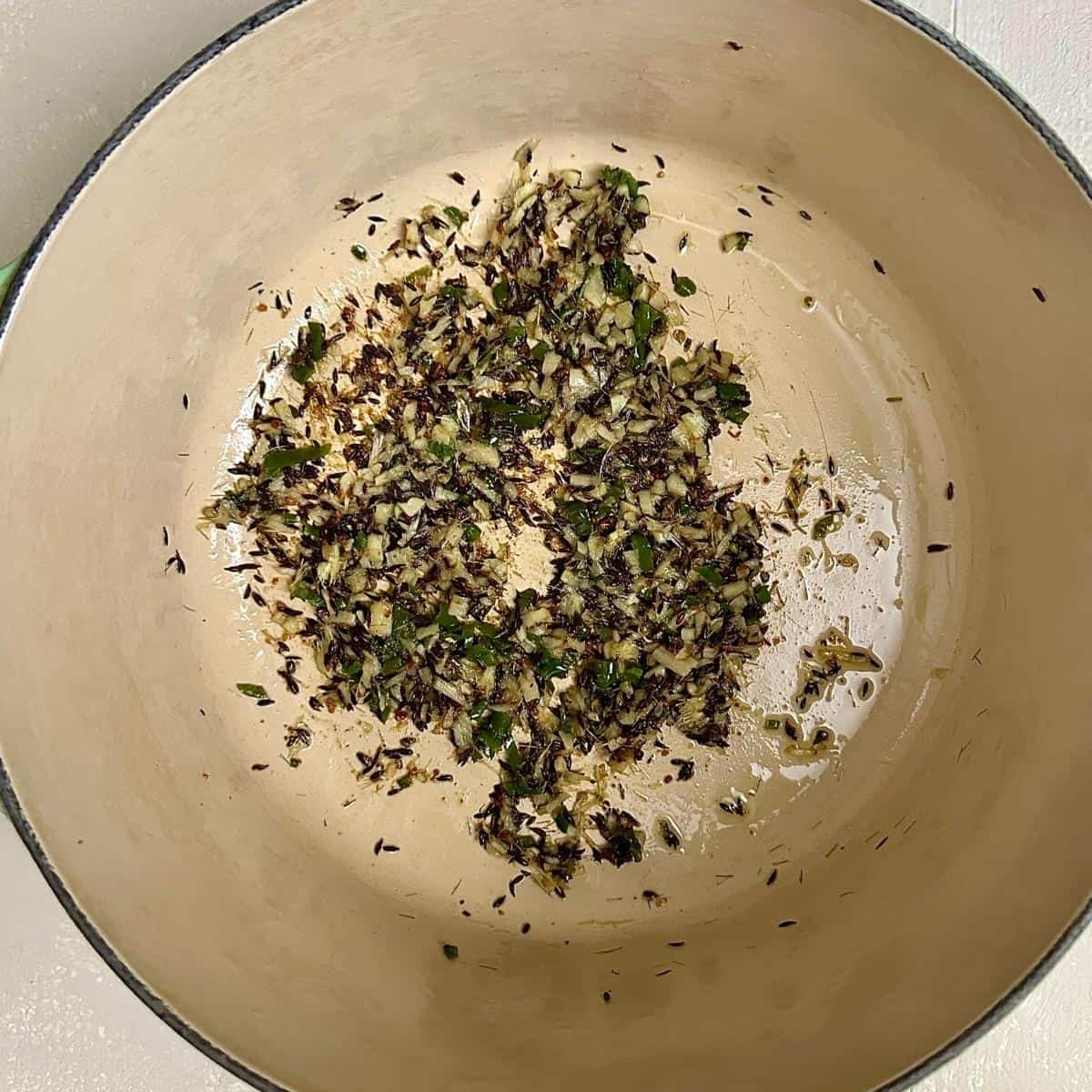 Bloomed spices: cumin seeds, asafoetida, ginger and green chillies in a large green pot, for pinwheel samosa