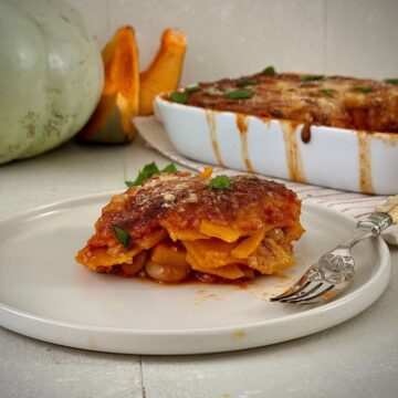 A slice of pumpkin casserole with butterbeans on a white plate. A casserole dish and crown prince pumpkin sit in the background.