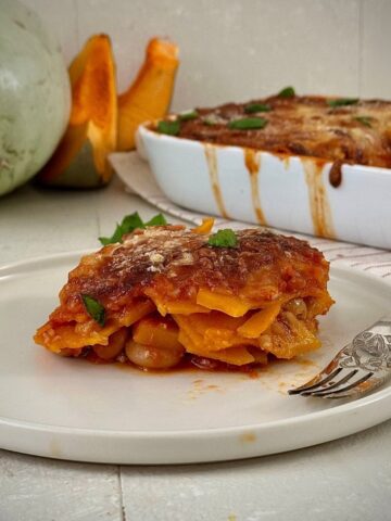 A slice of pumpkin casserole with butterbeans on a white plate. A casserole dish and crown prince pumpkin sit in the background.