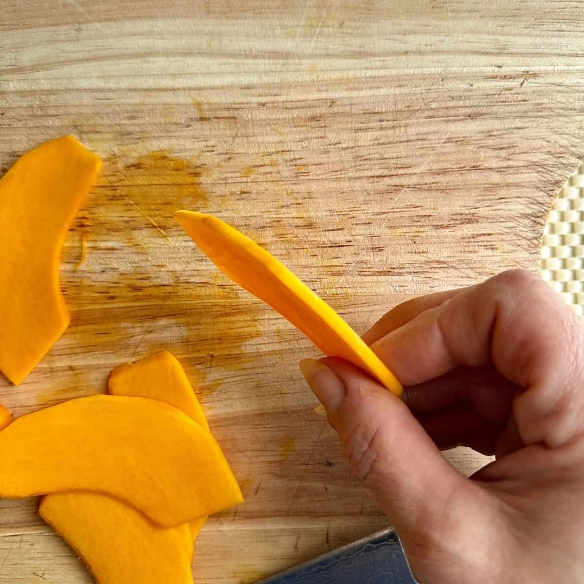 close up of a hand holding a thin slice of crown prince pumpkin to show the thickness of the slice. More slices sit on a chopping board underneath the hand.
