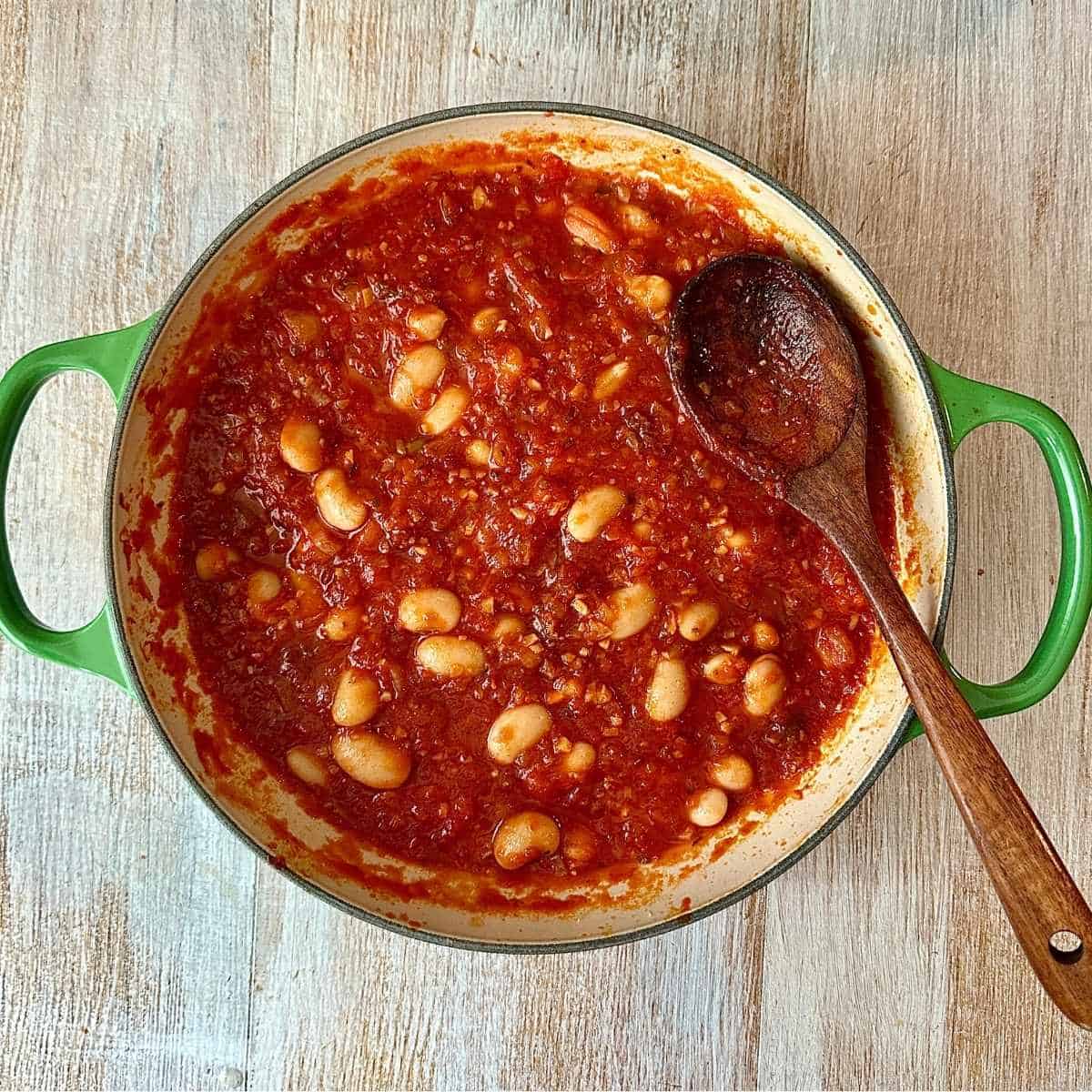 A large green p containing tomato passata and and butterbeans, for a pumpkin casserole. A wooden spoon sits in the pan.