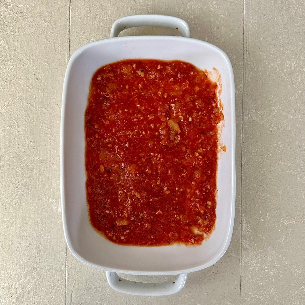 A white casserole dish containing a layer of tomato passata and butterbean sauce for a pumpkin casserole.