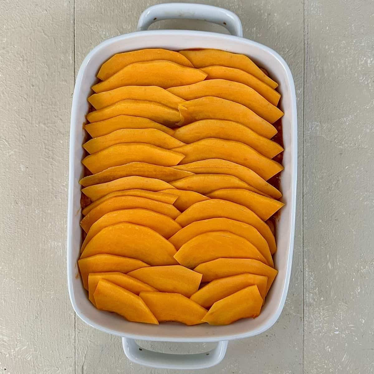 A white casserole dish containing a layer of thinly sliced pumpkin on top of a layer of butterbean and passata sauce. For a pumpkin casserole.