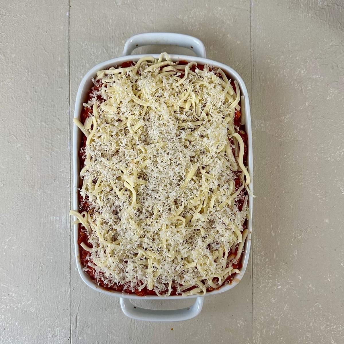 A white casserole dish containing pumpkin casserole. A layer of grated mozzarella and parmesan sits on top, ready to go into the oven to bake.