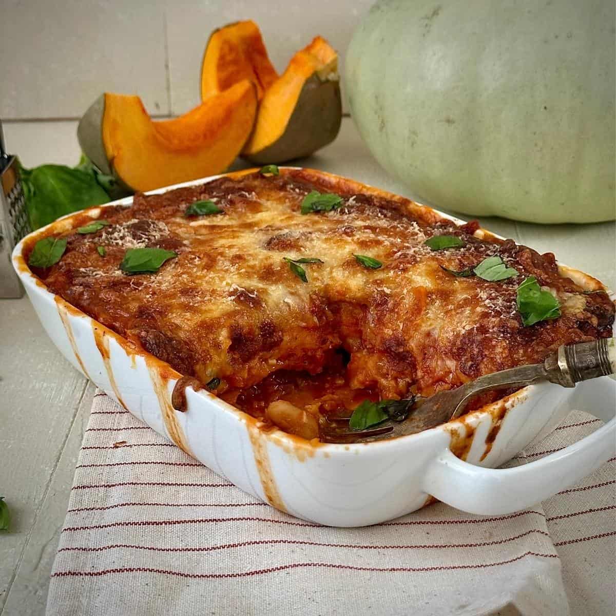 A pumpkin and butterbean casserole with a melted cheese topping in a white casserole dish. A crown prince pumpkin sits in the background.
