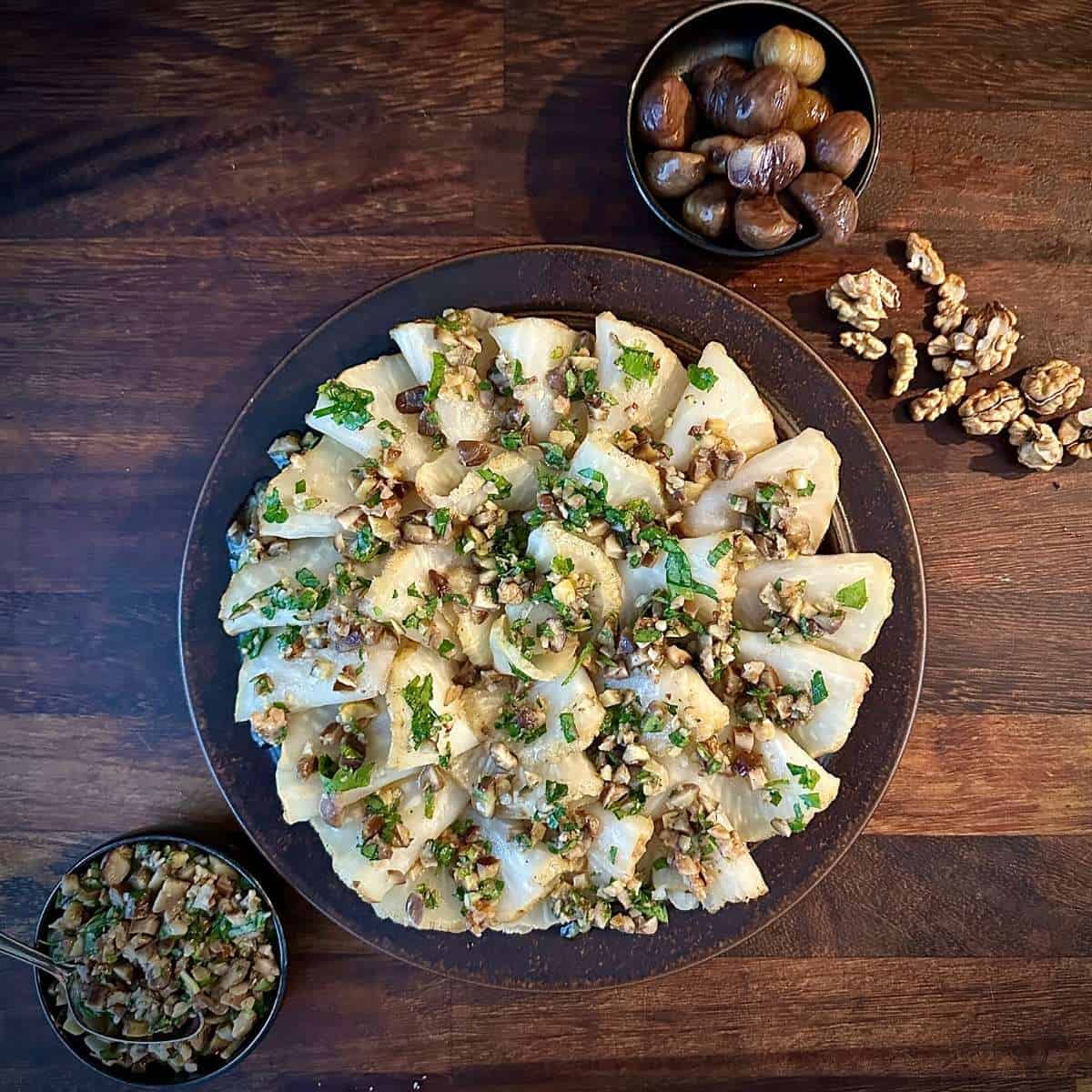 A brown plate containing roast celeriac, carpaccio-style, topped with orange, chestnut and walnut dressing. A small bowl of chestnuts sits to the top right with walnuts scattered next to it, and a small bowel of the dressing sits to the bottom left.