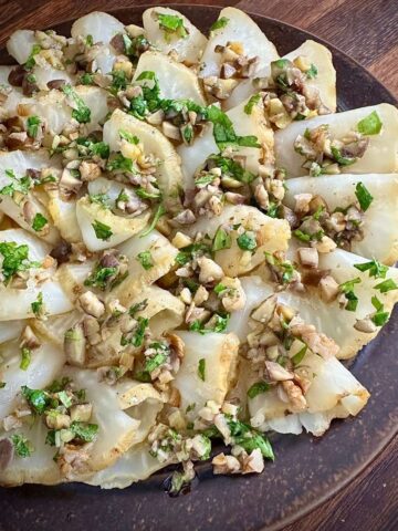 Close up of a plate of carpaccio-style roasted celeriac with an orange chestnut and walnut dressing garnished with parsley.
