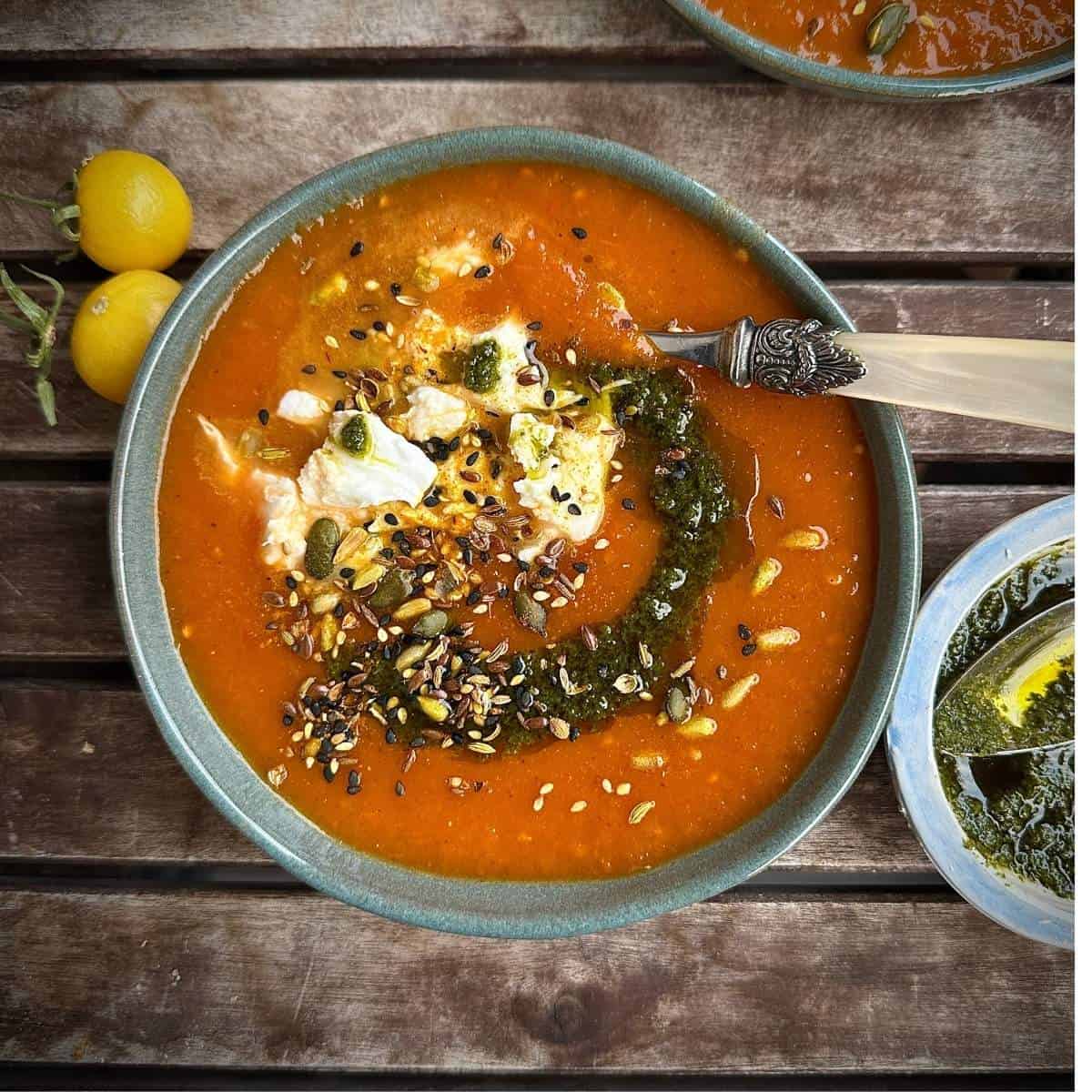 A bowl containing roasted tomato, leek and red pepper soup with torn pieces of mozzarella and garnished with a herb sauce and mixed seeds.