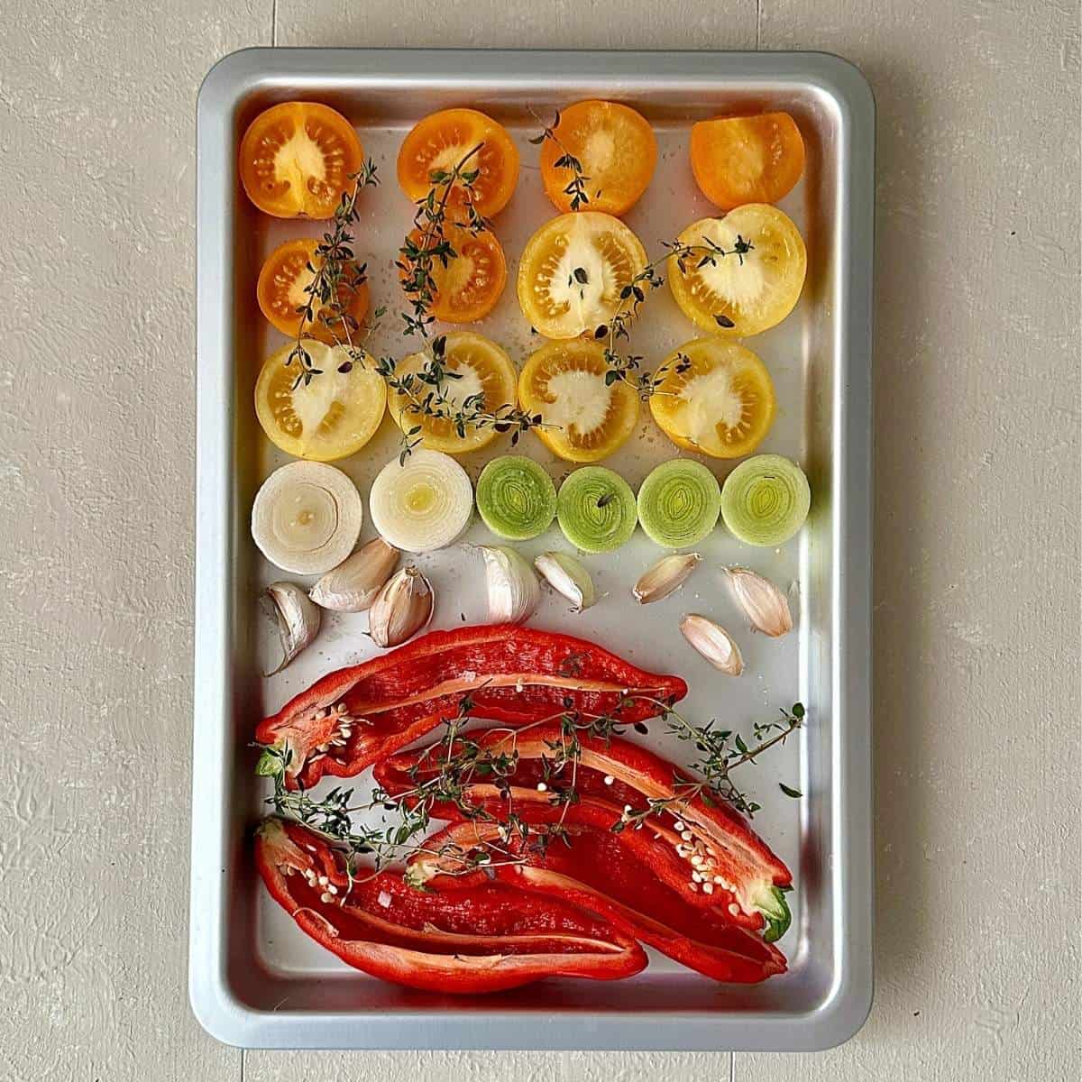 A sheet pan (baking tray) containing sliced rep peppers, sliced tomatoes, sliced leek and garlic, drizzled with olive oil and salt and cover with sprigs of thyme. Ready for roasting.