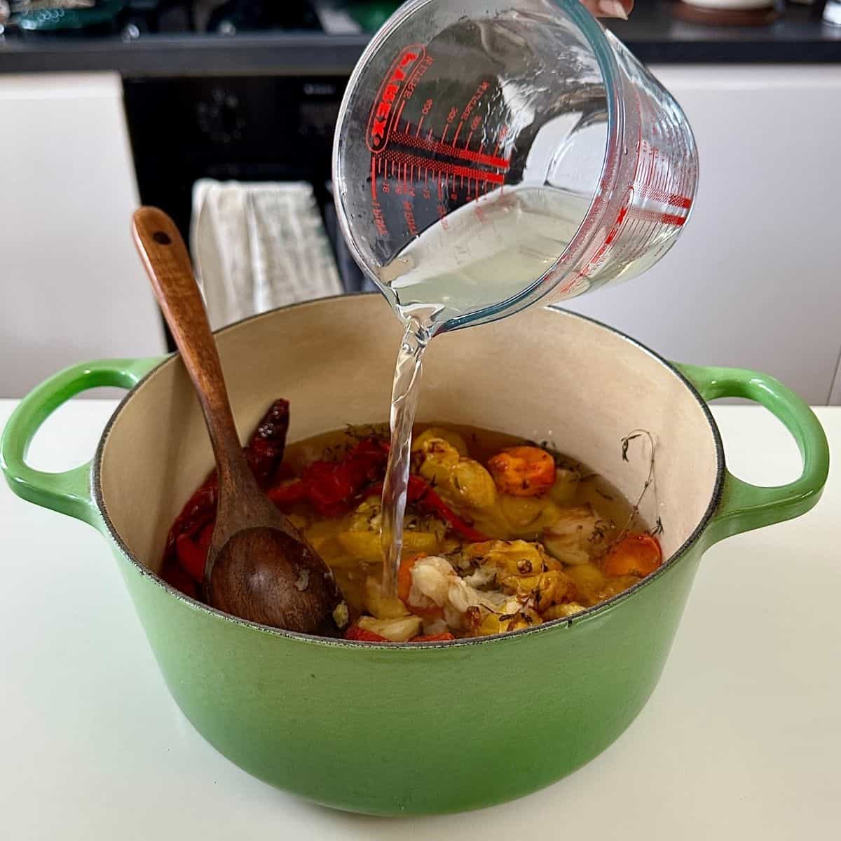 Pouring vegetable stock from a glass measuring jug into a large green saucepan contains roasted vegetables for roasted tomato, leek and red pepper soup.