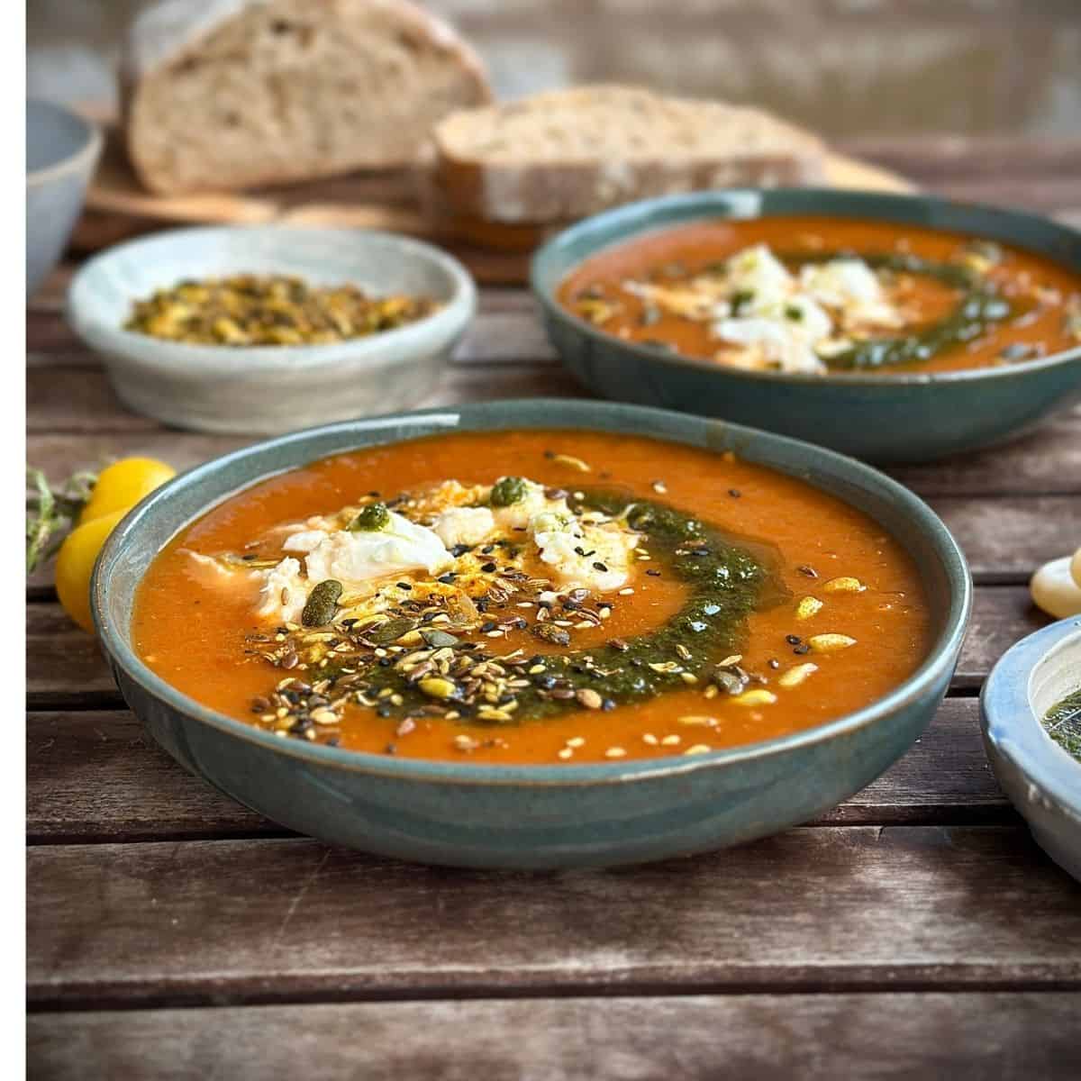 A green ceramic bowl containing tomato, leek and red pepper soup garnished with torn mozzarella, pesto and seeds.