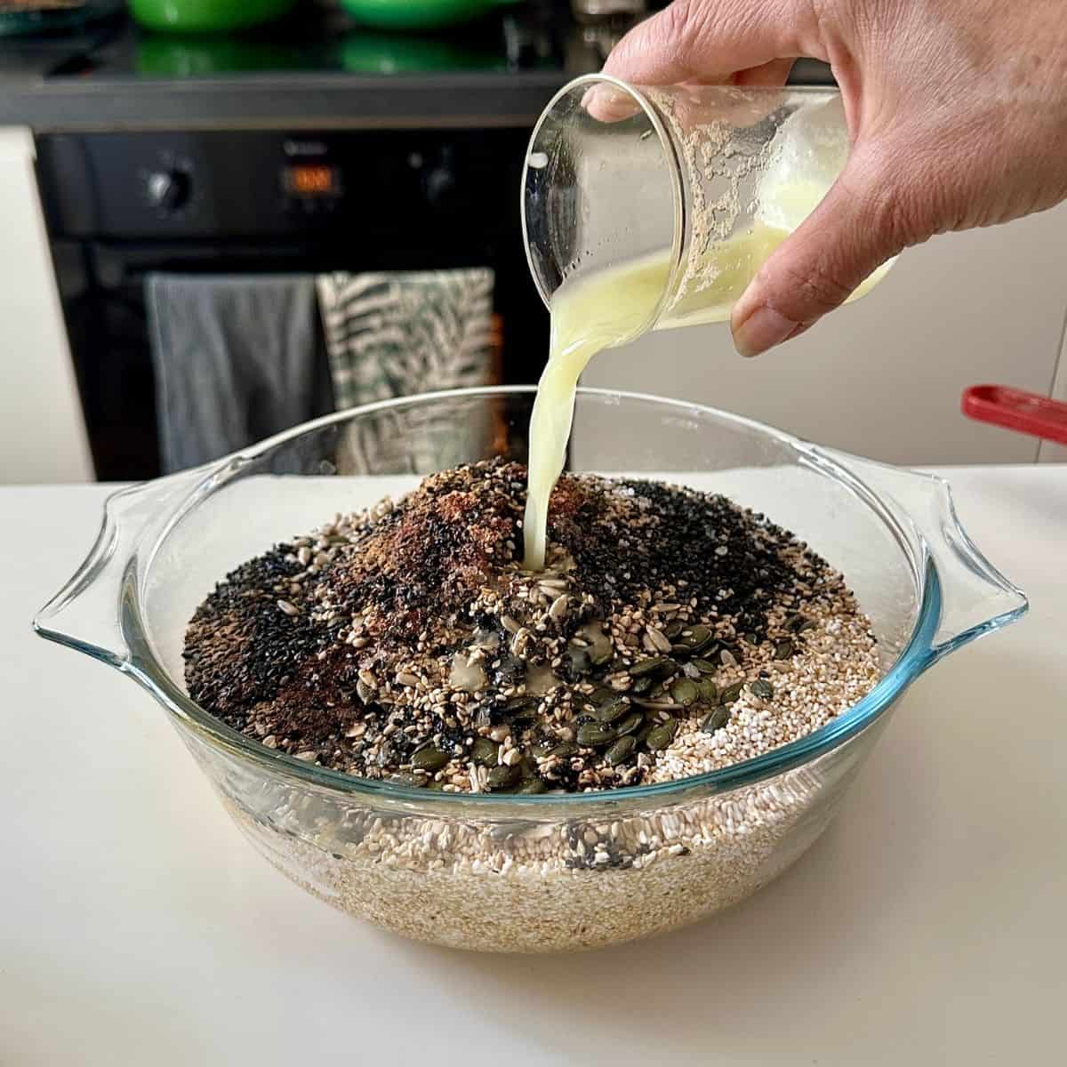 A hand pouring fresh apple juice from a glass beaker into a large glass bowl containing mixed dry ingredients of seeds, nuts, coconut, cinnamon and salt.