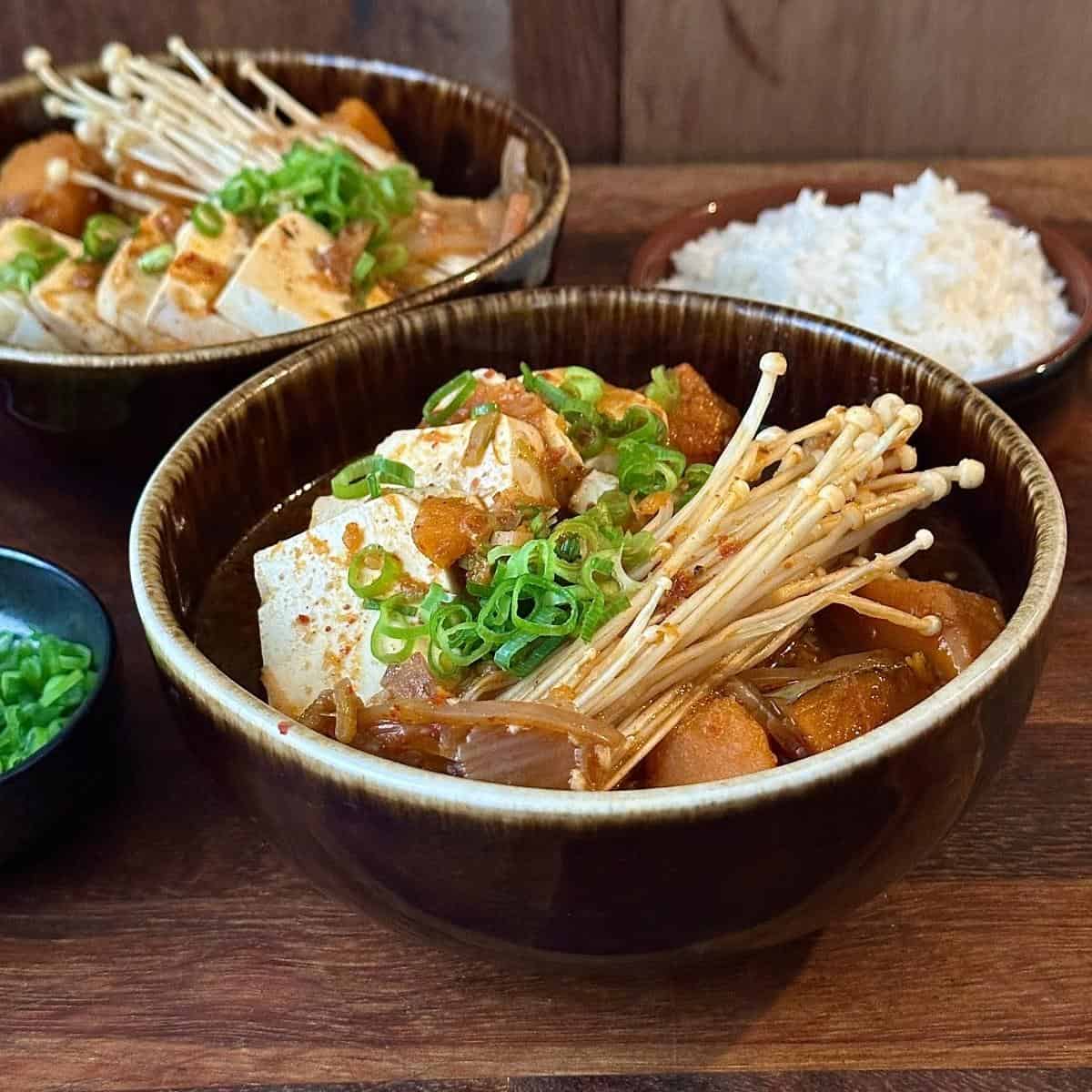 A brown ceramic bowl containing veggie kimchi jjigae. A small bowl of chopped spring onion garnish sits to the left and a bowl of steamed rice and another bowl of jjigae sit in the background.