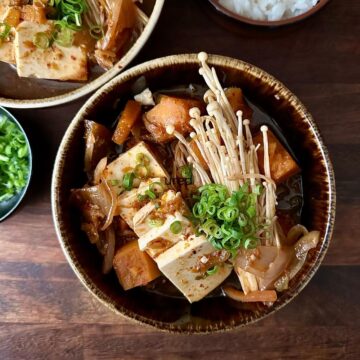 A bowl of veggie kimchi jjigae with tofu slices, brown prince pumpkin and enoki mushrooms in a broth, garnished with spring onions.