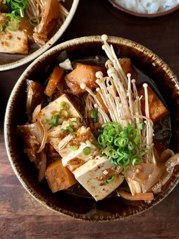 A bowl of veggie kimchi jjigae with tofu slices, brown prince pumpkin and enoki mushrooms in a broth, garnished with spring onions.