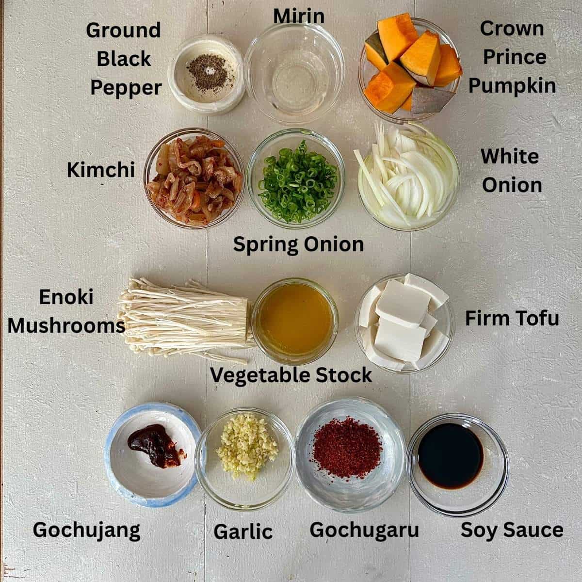 Prepared ingredients for veggie kimchi jjigae. The ingredients are in small glass and ceramic dishes. The ingredients are annotated.