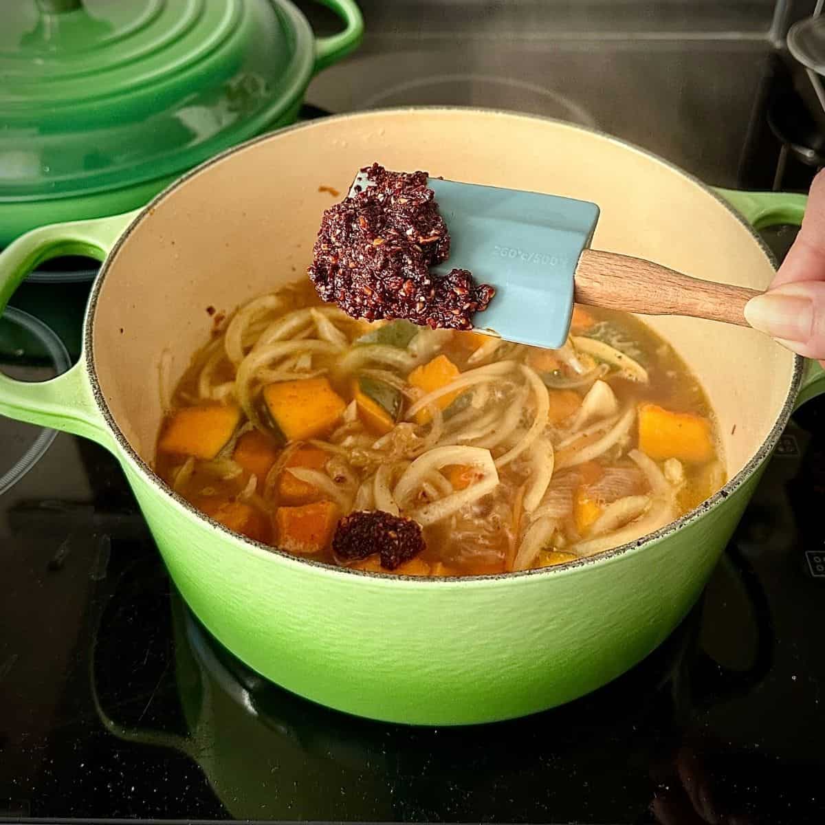 A large green pot containing veggie kimchi jjigae ingredients. A spatula is held over the pot with jjigae base mix on it, ready to stir into the other ingredients.