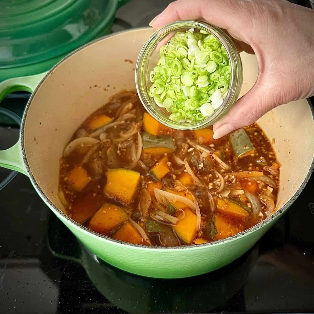 A large green pot contains ingredients for veggie kimchi jjigae. A hand holds a small glass dish containing finely chopped spring onion over the pot, ready to tip the spring onion into the pot.