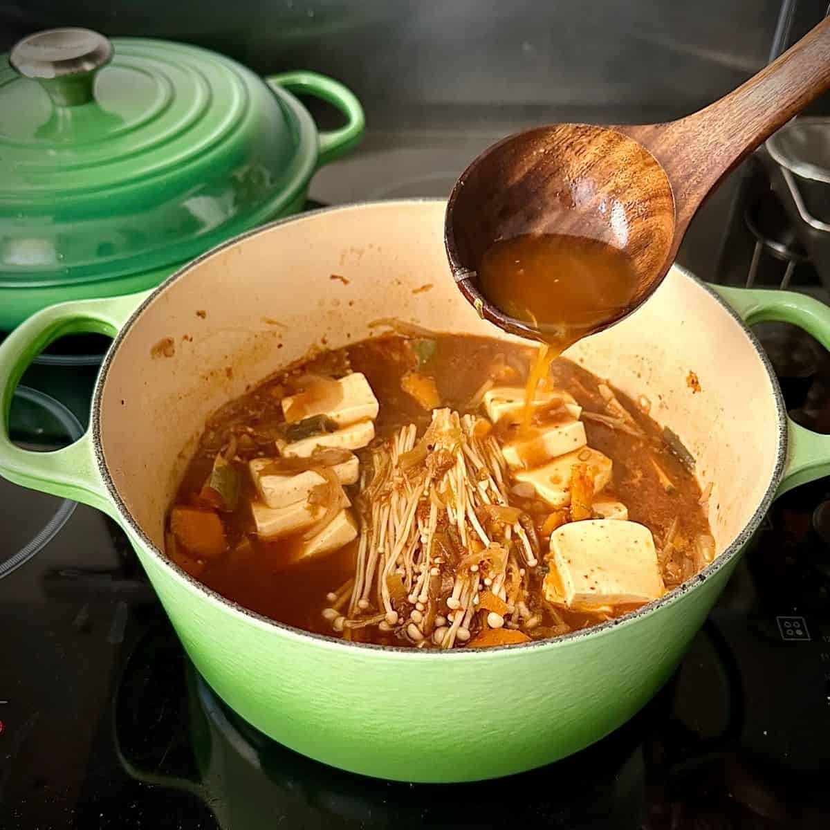 A large green pot containing veggie kimchi jjigae with tofu slabs and enoki mushrooms added. A wooden spoon is held over the pot, pouring hot stock over the mushrooms.
